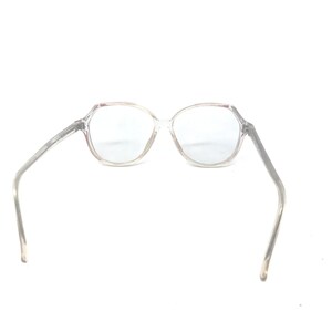May include: Clear, octagonal-shaped eyeglasses with a transparent frame and temples. The glasses are set against a white background, showcasing their design and clarity. The lenses appear to be clear.
