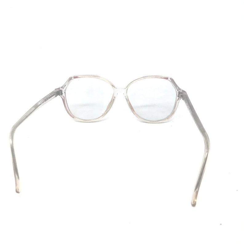 May include: Clear, octagonal-shaped eyeglasses with a transparent frame and temples. The glasses are set against a white background, showcasing their design and clarity. The lenses appear to be clear.