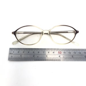 May include: Oval-shaped eyeglasses with a gradient brown and clear frame. The glasses are approximately 5.5 inches long. A metal ruler is placed below the glasses for measurement.
