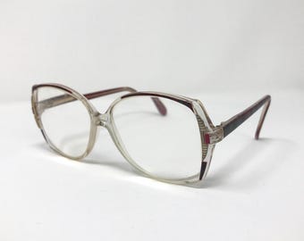 Könnte beinhalten: Klarer Kunststoff-Brillengestell mit burgunderfarbenem Rand. Die Brille hat eine quadratische Form mit abgerundeten Ecken.