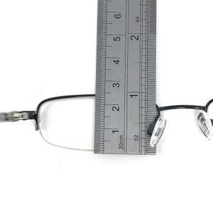 K&ouml;nnte beinhalten: Eine Brille mit schwarzem Rahmen und klaren Gl&auml;sern wird mit einem Lineal gemessen. Die Brille hat eine rechteckige Form und transparente Nasenpads. Das Lineal zeigt eine Messung von etwa 7,6 cm an.