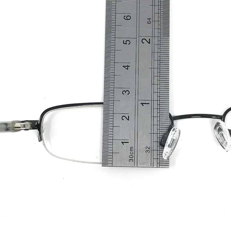 K&ouml;nnte beinhalten: Eine Brille mit schwarzem Rahmen und klaren Gl&auml;sern wird mit einem Lineal gemessen. Die Brille hat eine rechteckige Form und transparente Nasenpads. Das Lineal zeigt eine Messung von etwa 7,6 cm an.
