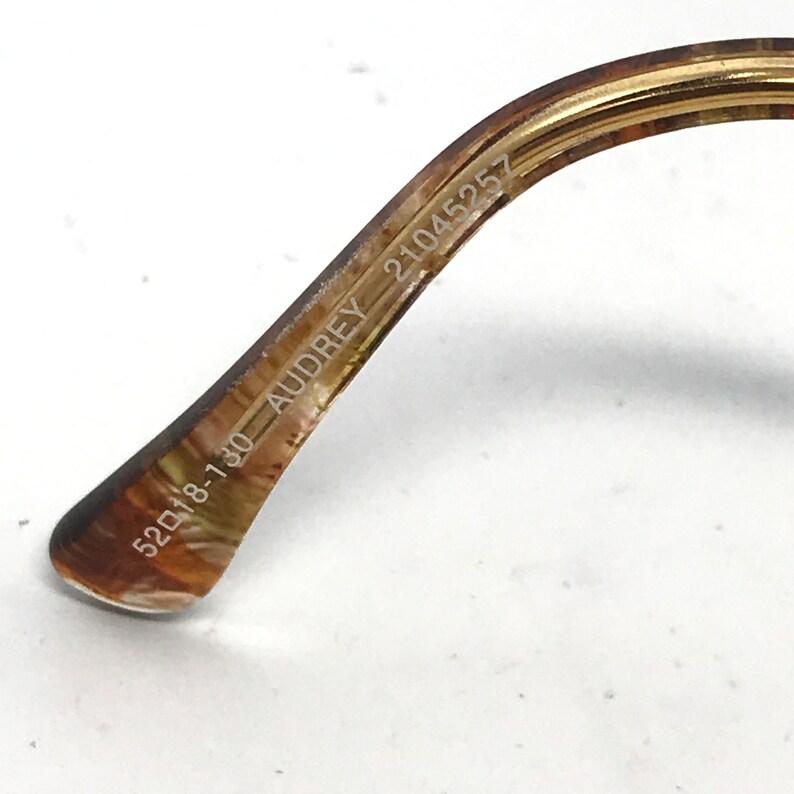 May include: Close-up of the temple arm of eyeglasses. The arm is a marbled brown and gold color. The text "52018-130 AUDREY 21045257" is printed on the arm.