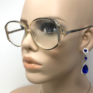 Puede incluir: Un par de gafas de montura dorada con lentes transparentes y un marco estampado en gris y negro. Las gafas las lleva puestas un maniquí con pendientes azules en forma de lágrima.