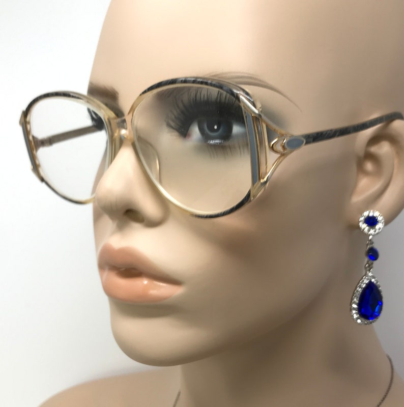 Puede incluir: Un par de gafas de montura dorada con lentes transparentes y un marco estampado en gris y negro. Las gafas las lleva puestas un maniqu&iacute; con pendientes azules en forma de l&aacute;grima.