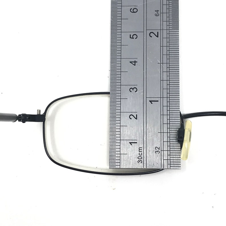 Puede incluir: Montura de gafas negra de forma rectangular, medida con una regla. La montura mide aproximadamente 7,6 cm de altura. Las gafas tienen una montura fina de metal negro y lentes transparentes.