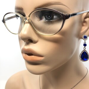 May include: A mannequin head wearing clear-framed eyeglasses and dangling blue and silver earrings. The earrings feature a cluster of small blue stones at the top, a small blue stone in the middle, and a large teardrop-shaped blue stone at the bottom, surrounded by clear stones.