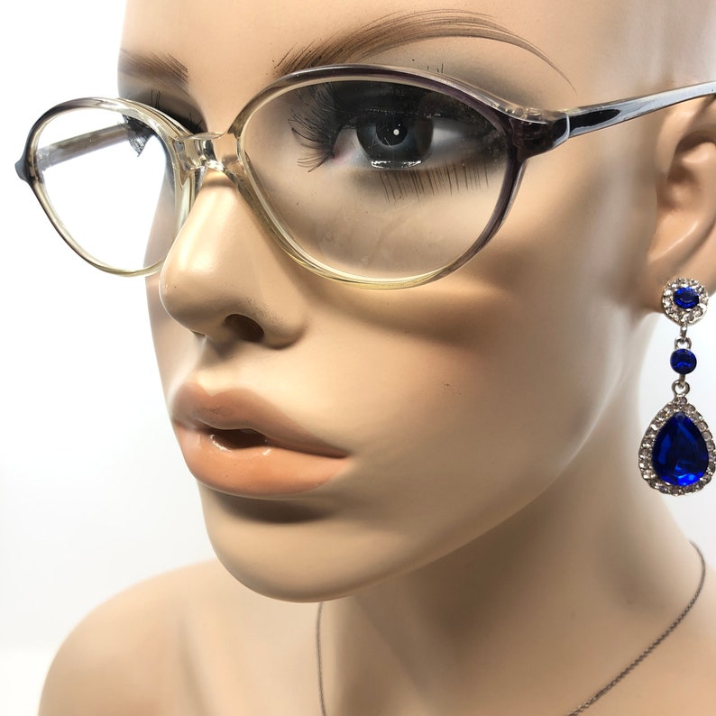 May include: A mannequin head wearing clear-framed eyeglasses and dangling blue and silver earrings. The earrings feature a cluster of small blue stones at the top, a small blue stone in the middle, and a large teardrop-shaped blue stone at the bottom, surrounded by clear stones.