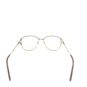 K&ouml;nnte beinhalten: Goldfarbene Brille mit ovalen Gl&auml;sern und schlanken B&uuml;geln. Die B&uuml;gel gehen in ein braunes Schildpattmuster in der N&auml;he der Ohren &uuml;ber. Die Brille ist vor einem wei&szlig;en Hintergrund platziert.