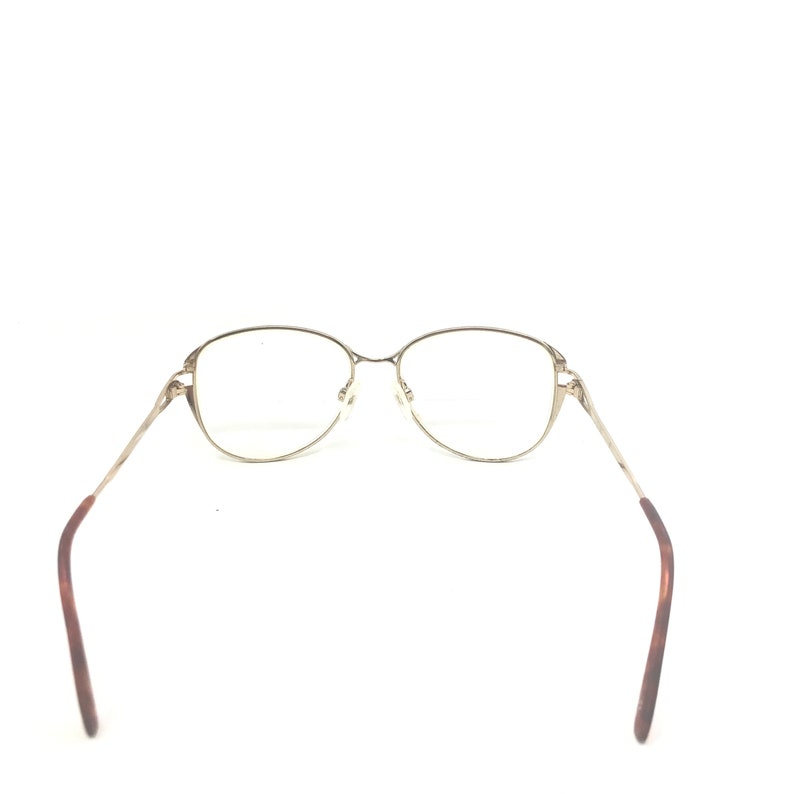 K&ouml;nnte beinhalten: Goldfarbene Brille mit ovalen Gl&auml;sern und schlanken B&uuml;geln. Die B&uuml;gel gehen in ein braunes Schildpattmuster in der N&auml;he der Ohren &uuml;ber. Die Brille ist vor einem wei&szlig;en Hintergrund platziert.