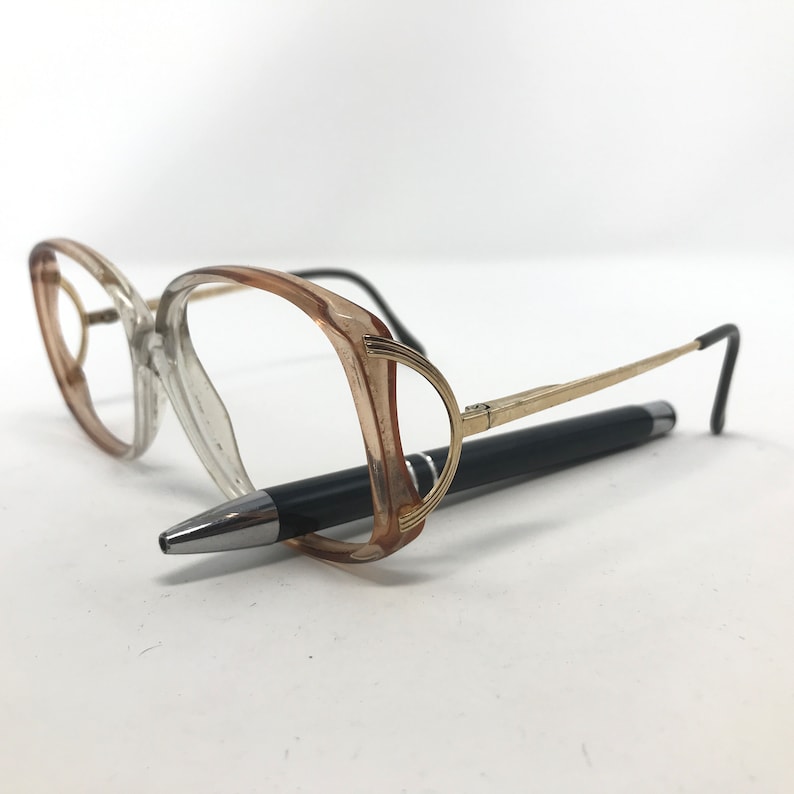 May include: A pair of eyeglasses with a clear and light brown frame, gold-colored arms, and black earpieces. A black pen rests beneath the glasses, creating a contrast against the white background.