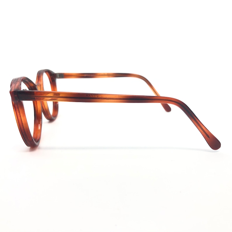May include: Brown and tan eyeglasses with a round frame and a striped pattern. The glasses have a classic design with a thin frame and temples. The lenses are clear, and the glasses are set against a white background.