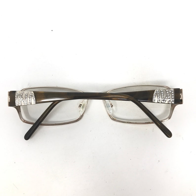 May include: Eyeglasses with a rectangular frame and clear lenses. The temples are dark brown with silver accents and rhinestones. The frame is a light brown color.