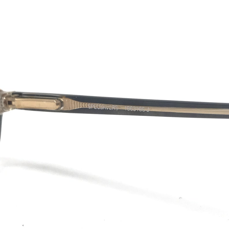 May include: Close-up of an eyeglasses arm with the text "SPECSAVERS 10631053 J". The arm is black with a gold-coloured accent near the hinge. The frame is a neutral colour.