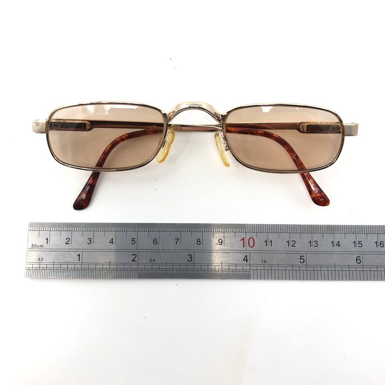 May include: Rectangular eyeglasses with brown tinted lenses and a gold-colored metal frame. The temples have a tortoiseshell pattern. The glasses are approximately 5.5 inches wide.