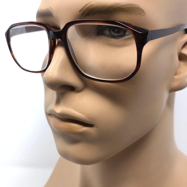 May include: A pair of eyeglasses with a dark brown, tortoiseshell-patterned frame. The glasses have a rectangular shape with rounded edges. The lenses are clear, and the temples are a solid dark brown. The glasses are displayed on a mannequin.