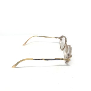 May include: A pair of vintage eyeglasses with a light brown frame and clear lenses. The temples are a light brown color with a gold-colored metal detail. The glasses are set against a white background.