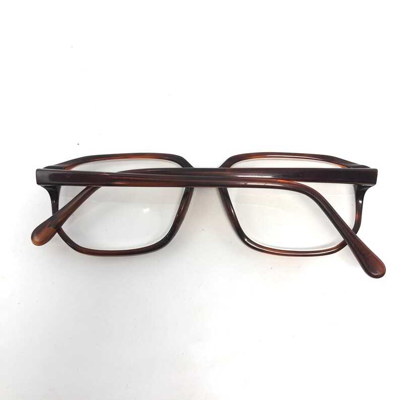 May include: Brown-framed eyeglasses with a rectangular shape. The glasses have a classic design with a dark brown color. The lenses are clear, and the frame is made of a glossy material. The arms of the glasses are curved for a comfortable fit.