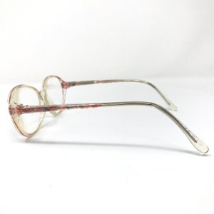 May include: Clear eyeglasses with a floral pattern on the frame and temples. The lenses are round, and the arms are thin and silver-toned. The glasses are viewed from the side against a white background.