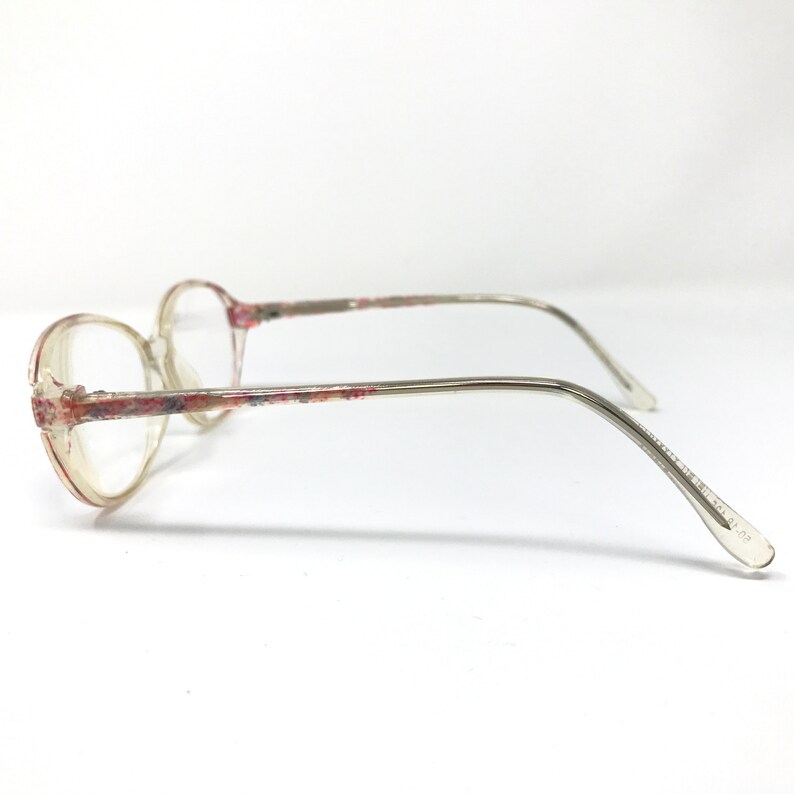 May include: Clear eyeglasses with a floral pattern on the frame and temples. The lenses are round, and the arms are thin and silver-toned. The glasses are viewed from the side against a white background.