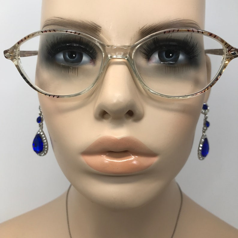 May include: Clear oval eyeglasses with a patterned top rim and temples. The glasses are set on a mannequin with blue teardrop earrings.
