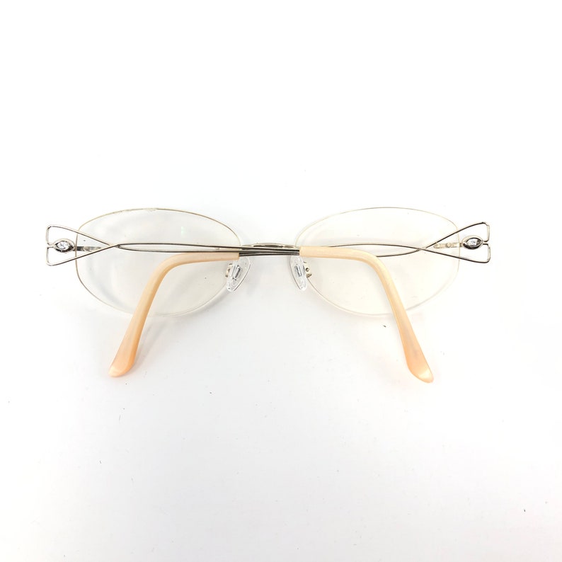 May include: Rimless eyeglasses with oval lenses and a silver-tone metal frame. The temples are a light peach color. The glasses have decorative accents at the outer edges of the lenses.
