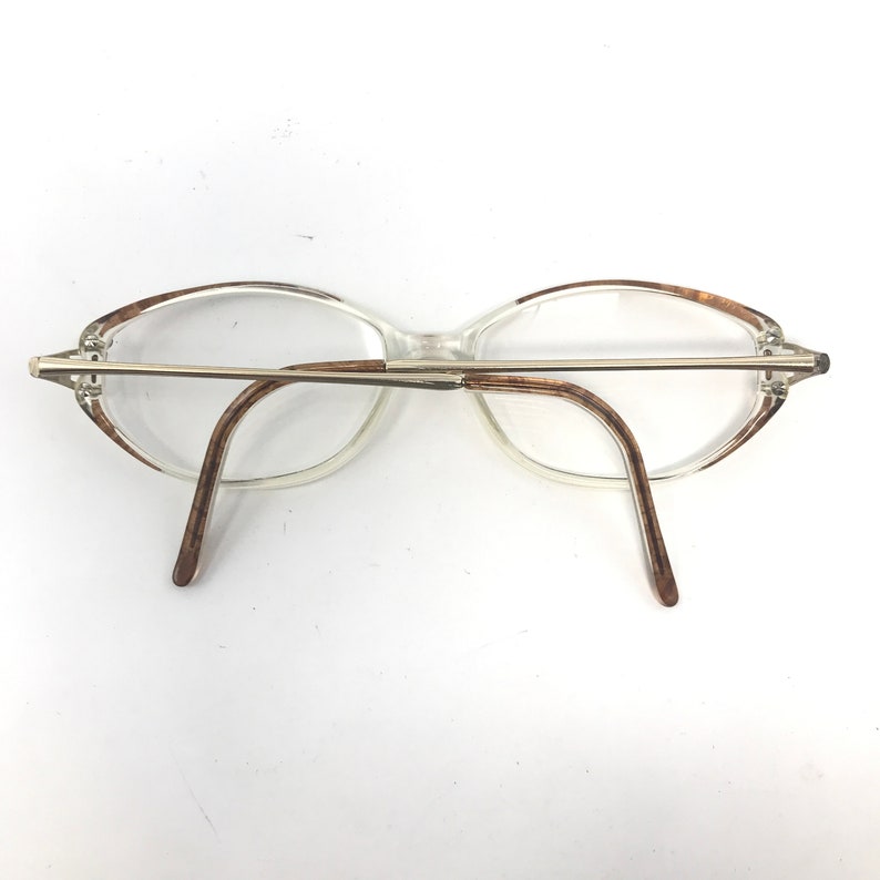 K&ouml;nnte beinhalten: Transparente Brille mit hellbraunen Akzenten an Rahmen und B&uuml;geln. Die Brille hat einen silberfarbenen Metallsteg und Scharniere. Die Gl&auml;ser sind klar, und das Design ist schlicht und klassisch.