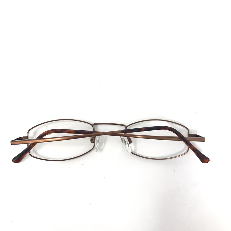 May include: A pair of eyeglasses with a thin, bronze-colored metal frame. The temples are a tortoiseshell pattern. The lenses are clear. The glasses are set against a white background.