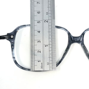 May include: Gray and white marbled eyeglasses with a ruler measuring the frame. The frame is approximately 13 cm wide. The glasses have a square shape and a metal hinge.