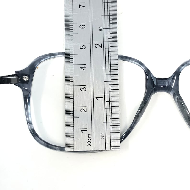 May include: Gray and white marbled eyeglasses with a ruler measuring the frame. The frame is approximately 13 cm wide. The glasses have a square shape and a metal hinge.