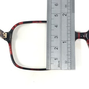 May include: Close-up of eyeglasses with a tortoiseshell pattern. A ruler is placed over the frame, measuring the width. The frame is black and red, with a rectangular shape. The hinge is silver.