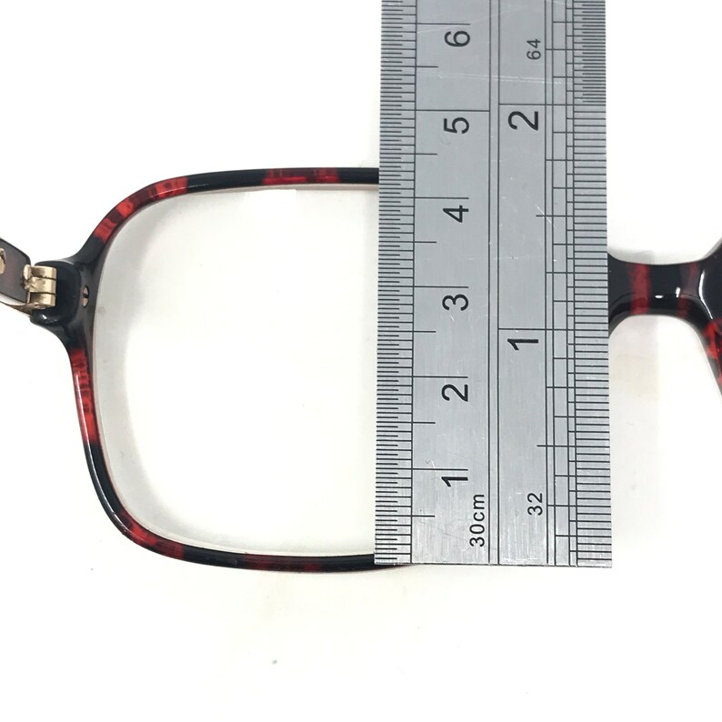May include: Close-up of eyeglasses with a tortoiseshell pattern. A ruler is placed over the frame, measuring the width. The frame is black and red, with a rectangular shape. The hinge is silver.