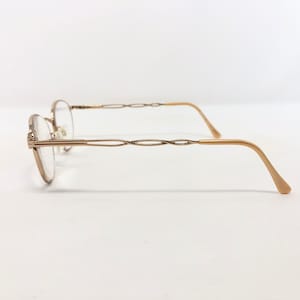 May include: A pair of gold-toned eyeglasses with oval lenses. The frames have a delicate, interwoven design on the temples and a curved, light-colored arm. The glasses are set against a plain white background.