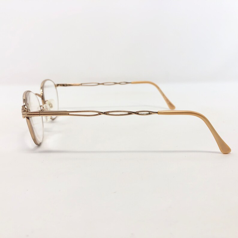 May include: A pair of gold-toned eyeglasses with oval lenses. The frames have a delicate, interwoven design on the temples and a curved, light-colored arm. The glasses are set against a plain white background.