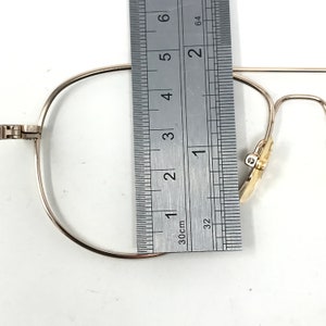 May include: Close-up of a pair of gold-colored eyeglasses with a ruler measuring the frame. The frame is approximately 2 inches wide. The glasses have a classic, minimalist design.