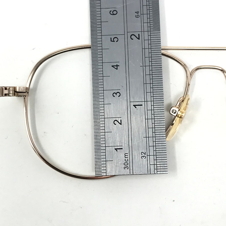 May include: Close-up of a pair of gold-colored eyeglasses with a ruler measuring the frame. The frame is approximately 2 inches wide. The glasses have a classic, minimalist design.