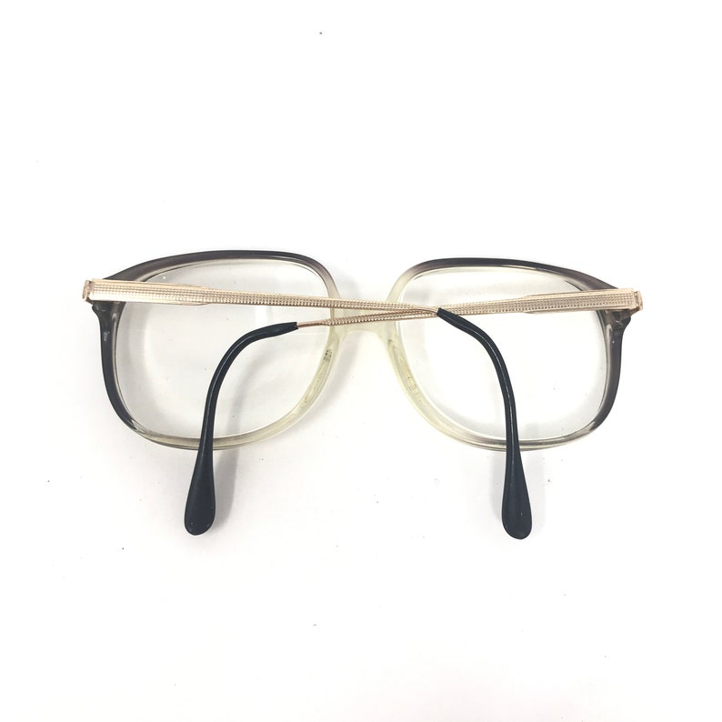 May include: A pair of eyeglasses with a clear frame that fades to a dark gray at the top. The glasses have a gold-colored bridge and black temples. The lenses are clear.