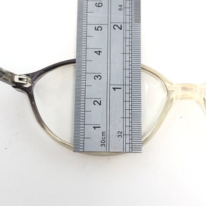 May include: Close-up of eyeglasses with a ruler measuring the frame. The glasses have a dark gray frame on one side and a clear, light frame on the other. The ruler shows measurements in inches and centimeters.
