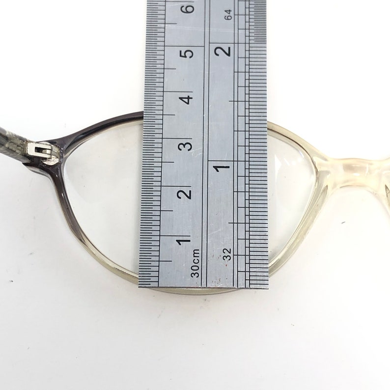 May include: Close-up of eyeglasses with a ruler measuring the frame. The glasses have a dark gray frame on one side and a clear, light frame on the other. The ruler shows measurements in inches and centimeters.