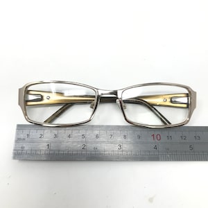 May include: Silver-framed eyeglasses with rectangular lenses and a gold-colored accent on the temples. The glasses are placed on a white surface next to a ruler, which measures up to 13 inches.