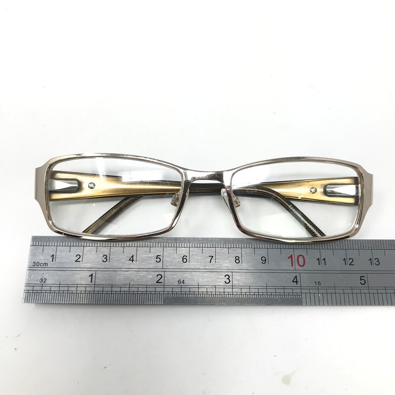 May include: Silver-framed eyeglasses with rectangular lenses and a gold-colored accent on the temples. The glasses are placed on a white surface next to a ruler, which measures up to 13 inches.