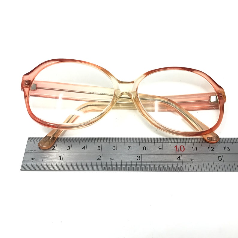 May include: A pair of large, round, peach-colored eyeglasses with a brown tint. The glasses are measured against a ruler, showing the width of the frame is approximately 10 centimeters.