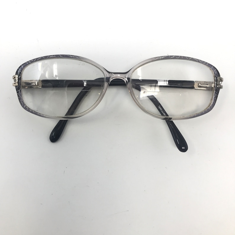 May include: Clear eyeglasses with oval lenses and a dark gray patterned frame. The temples are black, and the bridge is a dark color with silver accents. The glasses have the text "Spectre" on the temple.