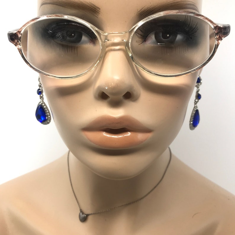 May include: A mannequin head wearing clear oval spectacles with patterned temples. The mannequin also has long eyelashes, blue teardrop earrings, and a small heart pendant necklace.