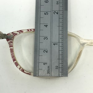 May include: Clear eyeglasses with a patterned frame in white and burgundy. A ruler is placed over the lens, measuring approximately 5 inches in height. The glasses have a classic, rectangular shape.