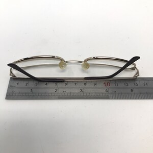 May include: Silver-framed eyeglasses with black temples and clear lenses. The glasses are positioned next to a ruler, with a measurement scale in inches. The overall design is minimalist and modern.