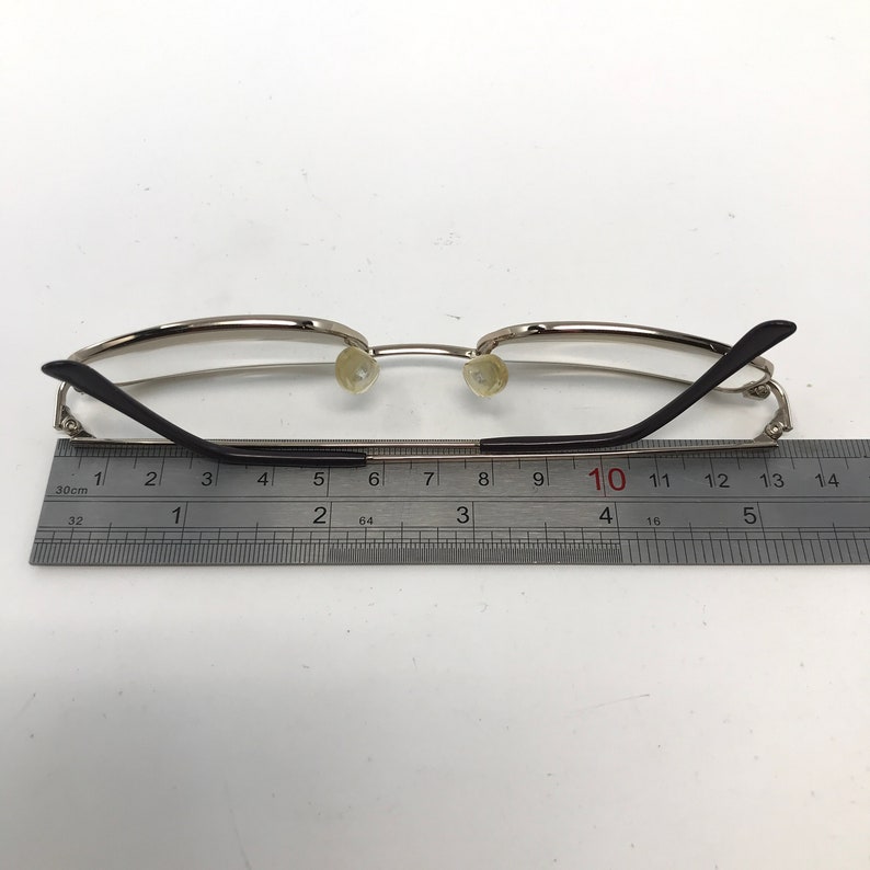 May include: Silver-framed eyeglasses with black temples and clear lenses. The glasses are positioned next to a ruler, with a measurement scale in inches. The overall design is minimalist and modern.