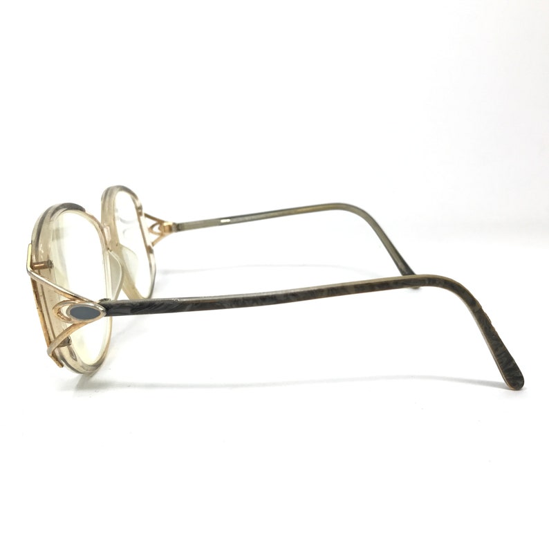 Puede incluir: Un par de gafas doradas con montura marr&oacute;n texturizada y lentes transparentes. La montura tiene un dise&ntilde;o &uacute;nico con un peque&ntilde;o detalle ovalado en el lateral.