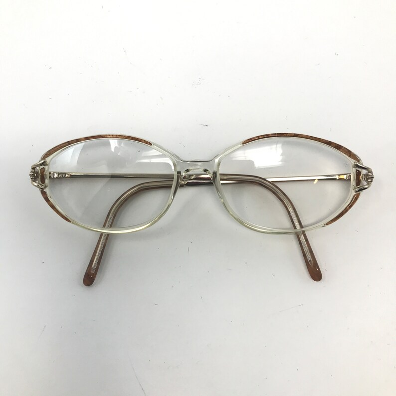 K&ouml;nnte beinhalten: Klare Brille mit ovalen Gl&auml;sern und hellbraunem Rand. Die Fassungen sind hell, durchscheinend mit silberfarbenen B&uuml;geln und braunen B&uuml;gelenden. Die Brille ist vor einem wei&szlig;en Hintergrund platziert.