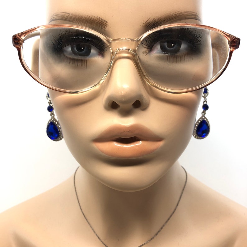 May include: Transparent-framed spectacles with a light brown tint, set against a mannequin's face. The glasses have a classic, slightly oversized shape. Dangling blue teardrop earrings with silver accents add a pop of colour, creating a vintage aesthetic.